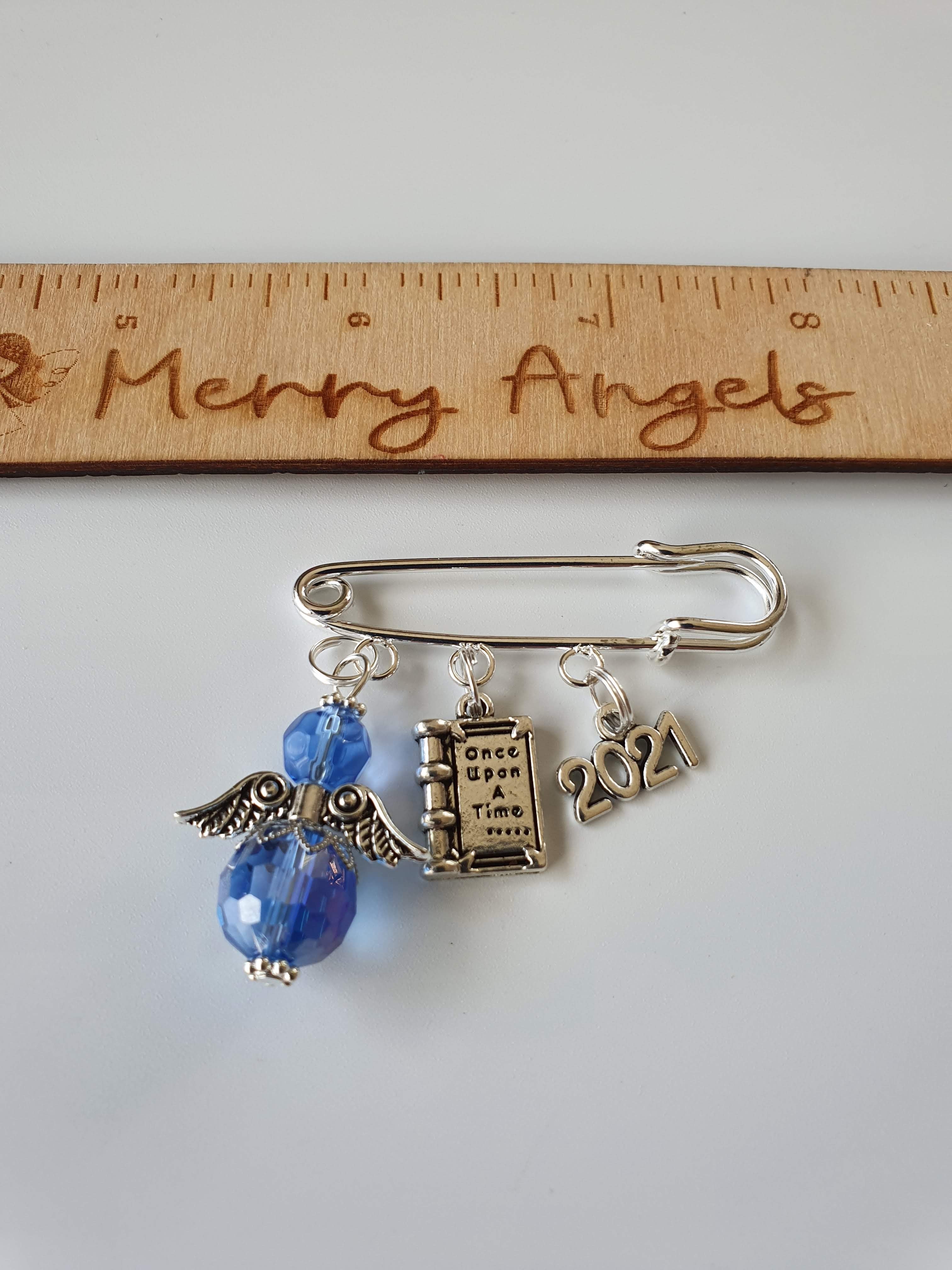 A silver pram pin with a blue angel with silver wings, a silver book charm with the words 'once upon a time' engraved on it, and a silver 2021 charm.