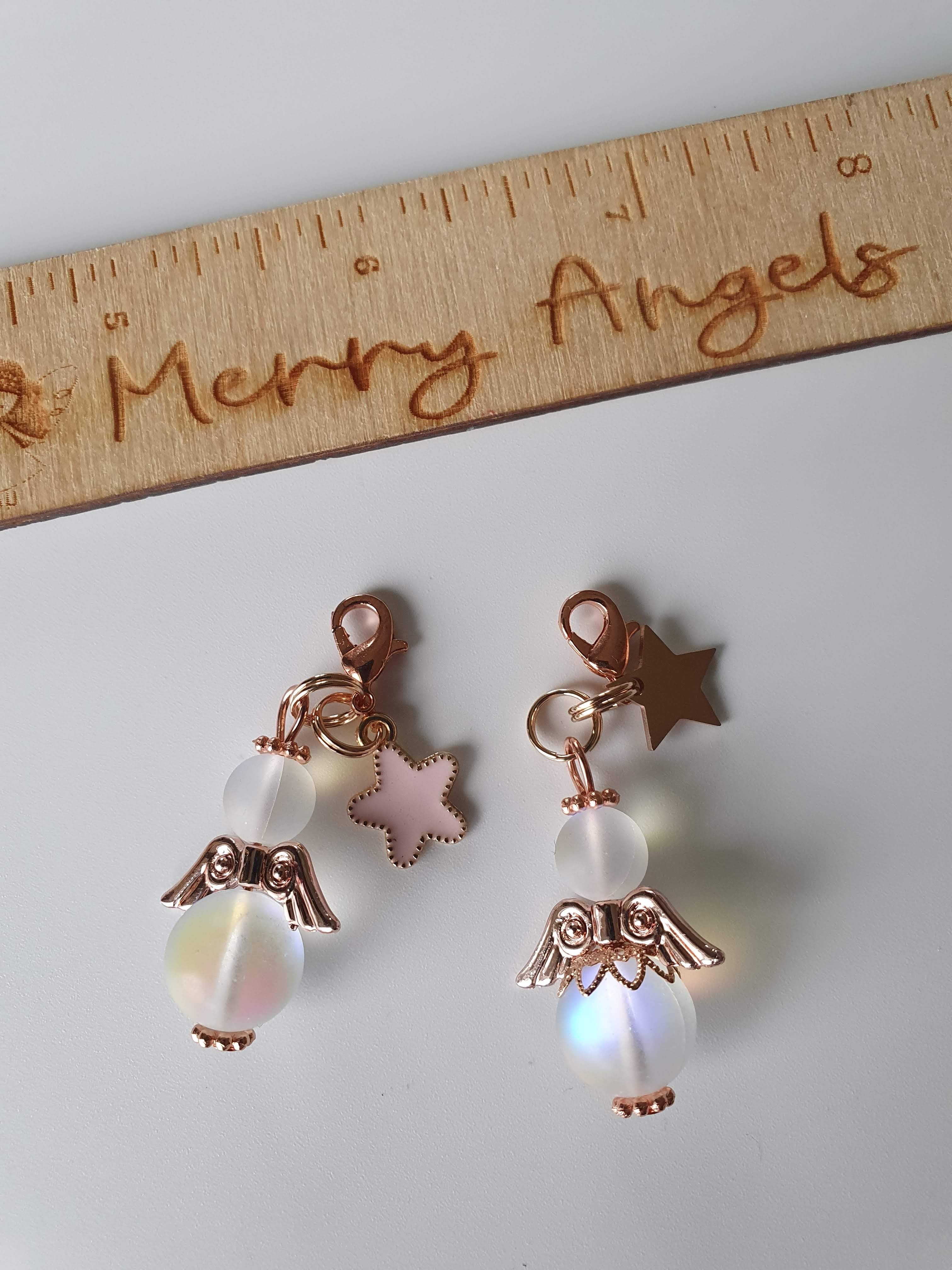 This is a picture of two angel hugs. The heads and bodies of the angels are white with rose gold wings and feet. Both angels have star charms attached.