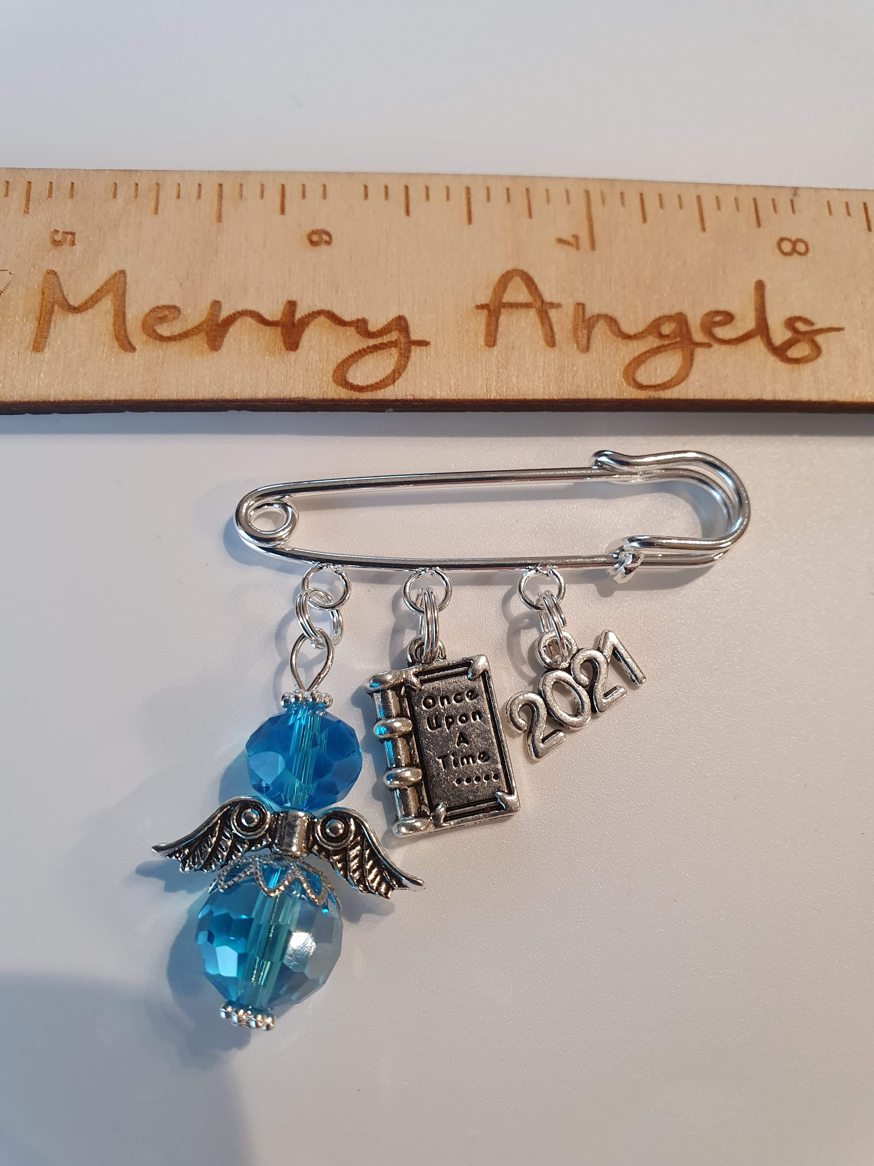 A silver pram pin with a blue angel with silver wings, a silver book charm with the words 'once upon a time' engraved on it, and a silver 2021 charm.