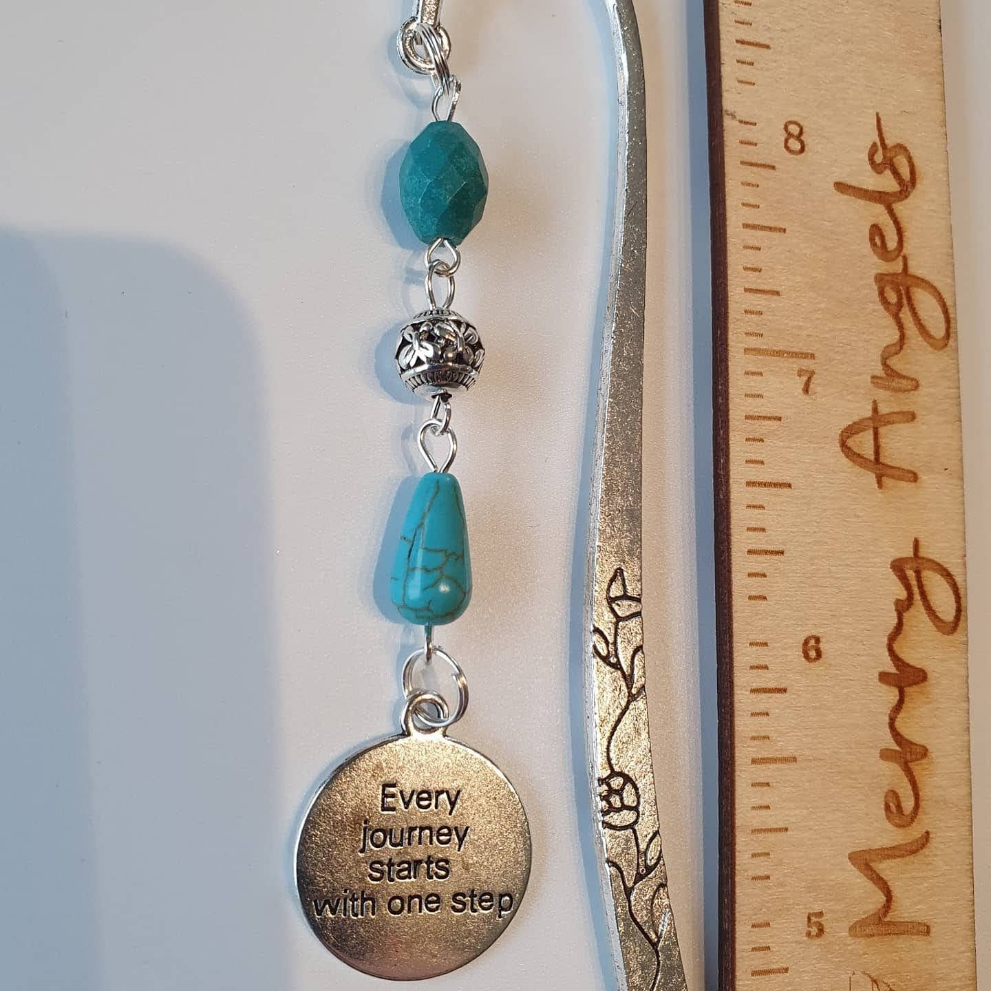 This is a picture of a silver bookmark with blue details and a silver circle charm with the words 'every journey starts with one step' engraved on it. 