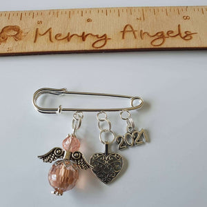 A silver plated pram pin with a peach angel with silver wings, a silver heart charm and a silver 2021 charm.