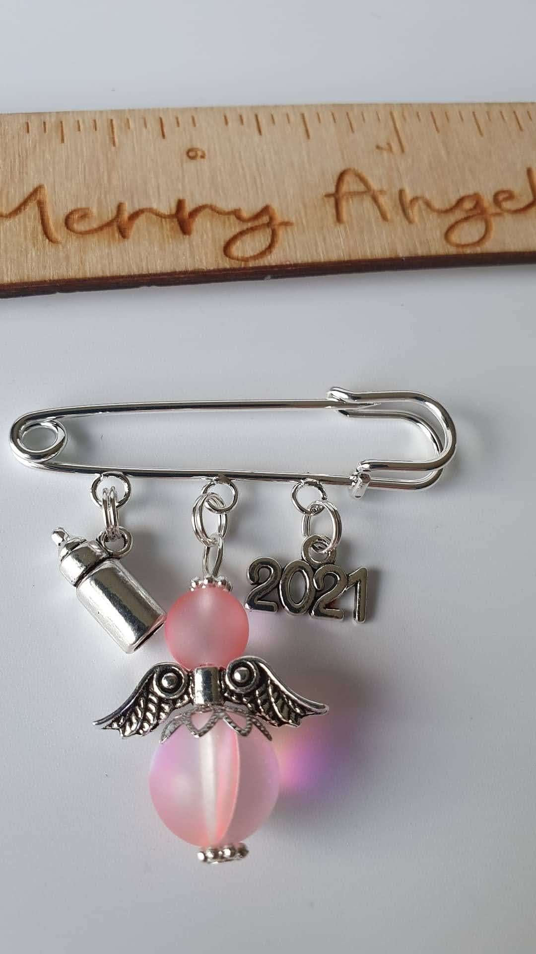 A silver plated pram pin with a silver bottle charm, a pink angel with silver wings, and a silver 2021 charm.