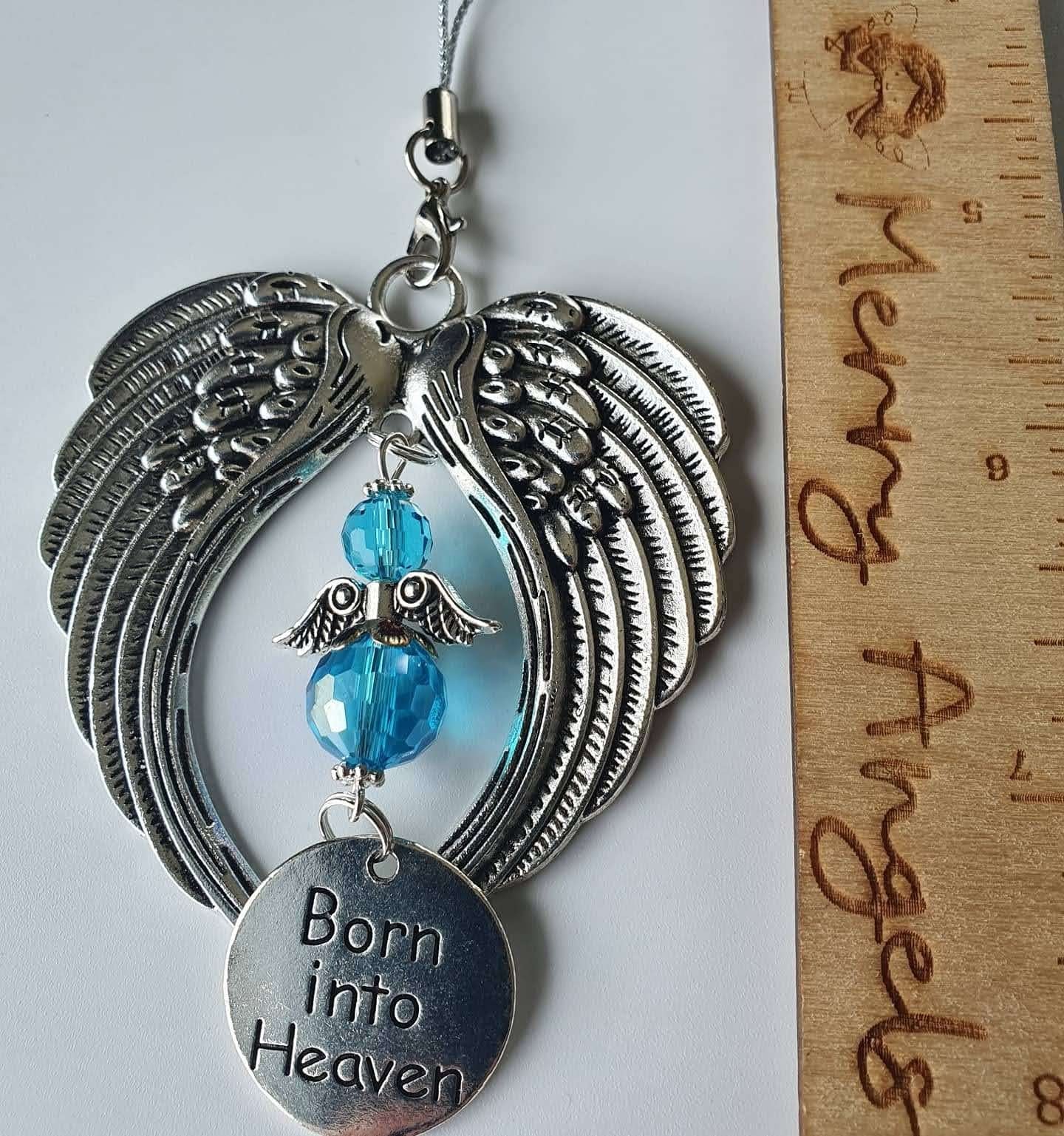 This is a picture of a silver angel wing hanging charm with a blue angel in the centre and a silver charm with the words 'born into heaven' engraved on it