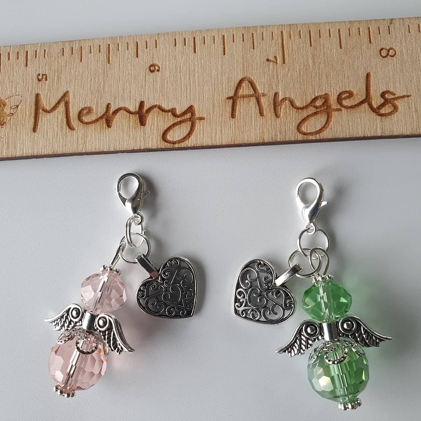 This is a picture of two angel hugs, one is a green colour and the other is a pale pink colour. Both have silver wings and feet and have silver heart charms attached. 
