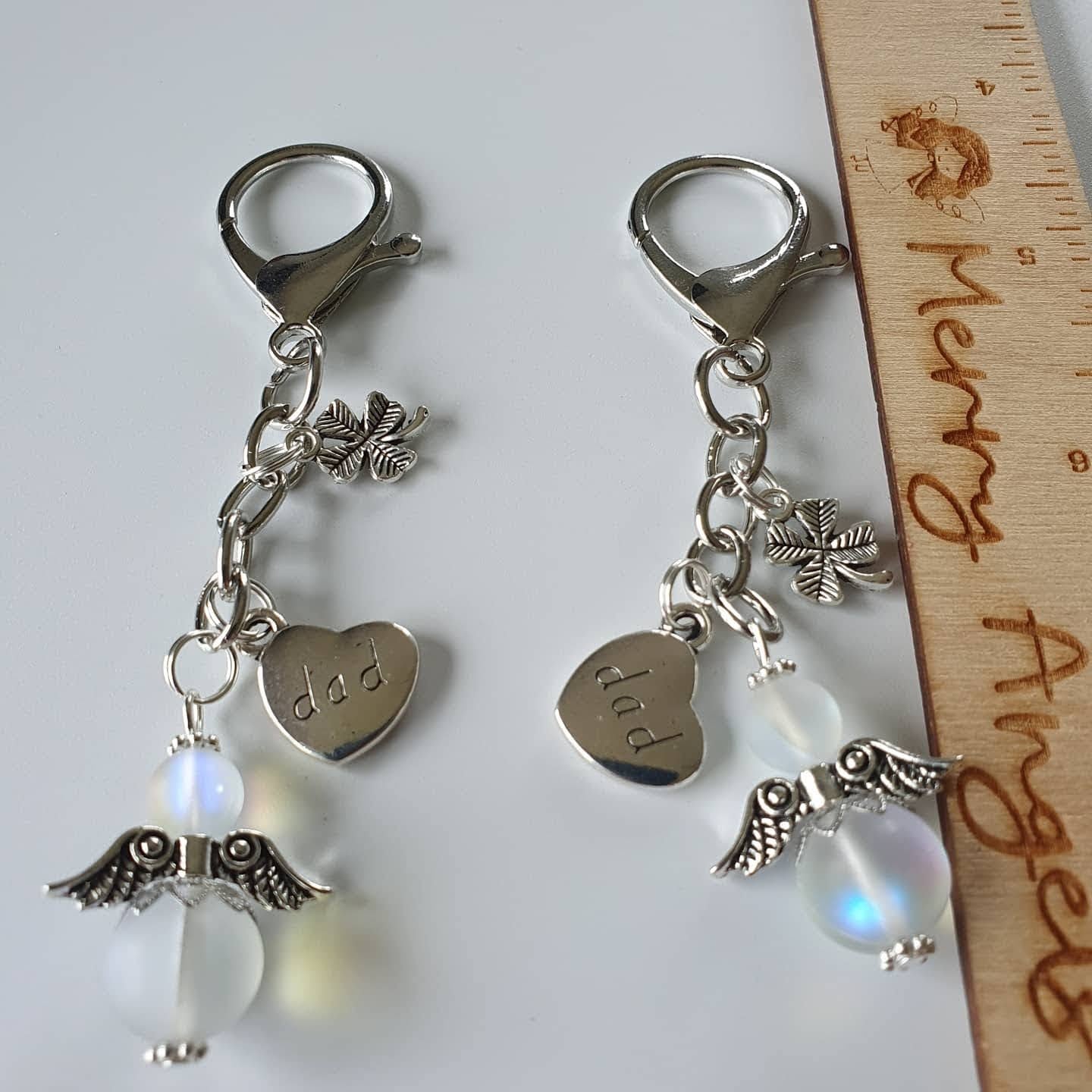 This is a picture of two silver angel keyrings, both keyrings have silver heart charms attached with the word 'dad' engraved on them and also silver shamrock charms
